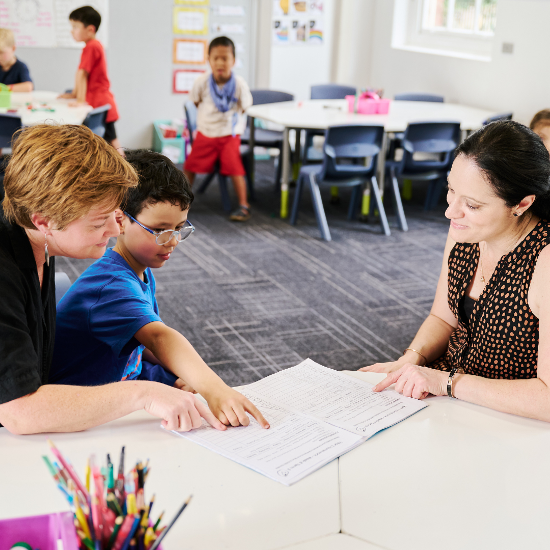 A photo of a young boy with dark hair wearing glasses sitting at a table in a classroom. On either side of him sit two women. The trio are looking and pointing at an open exercise book covered in handwritten text. Other school children can be seen in the background.