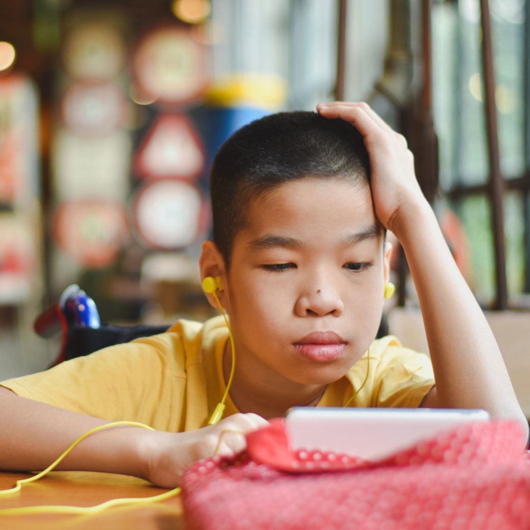 A photo of a young asian boy sitting at a table while watching something on his phone. He has a neutral facial expression and is wearing a yellow t-shirt and yellow earbuds.